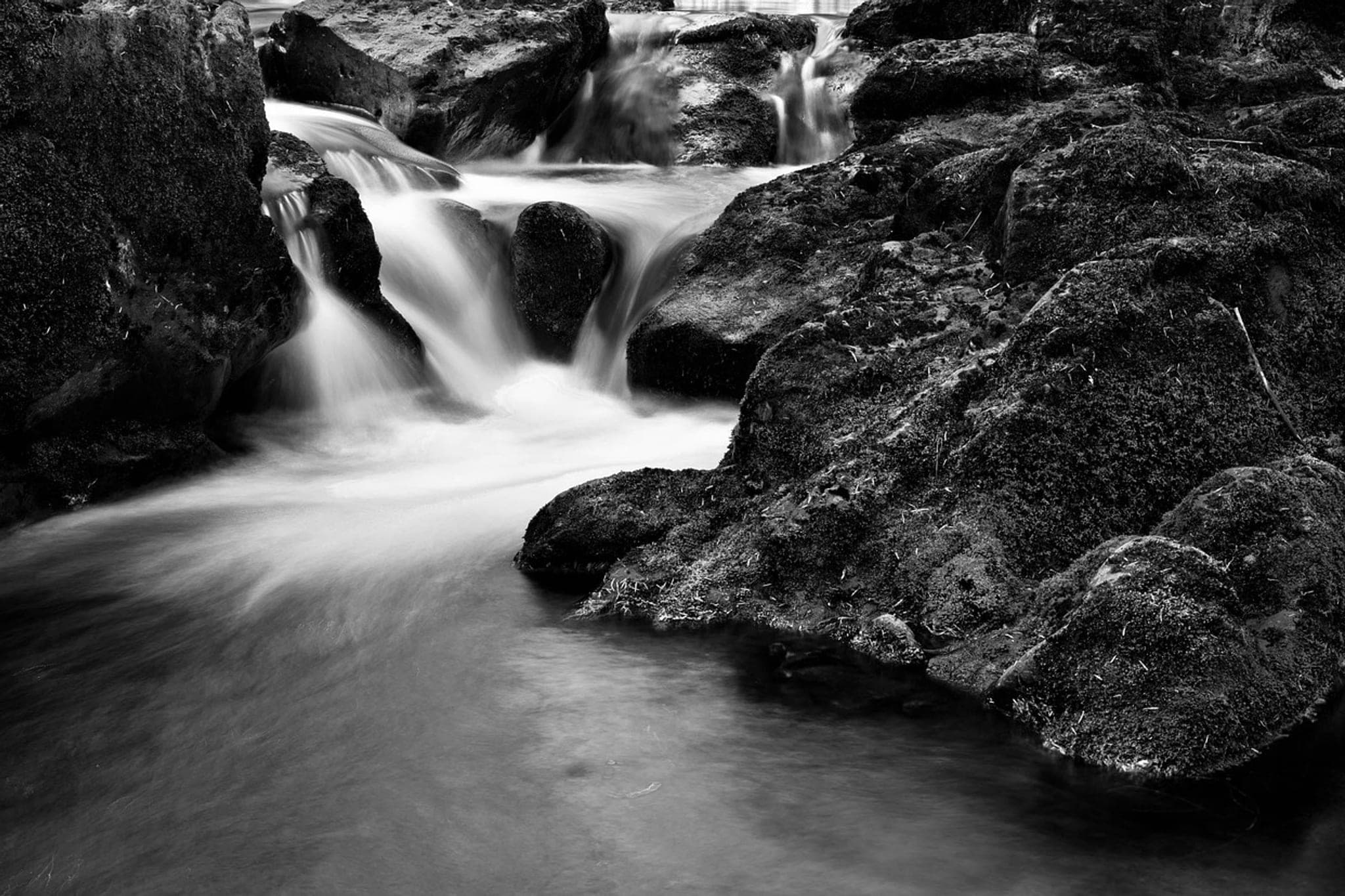 Water falling through a narrow gap between moss-covered boulders and collecting in a still pool below