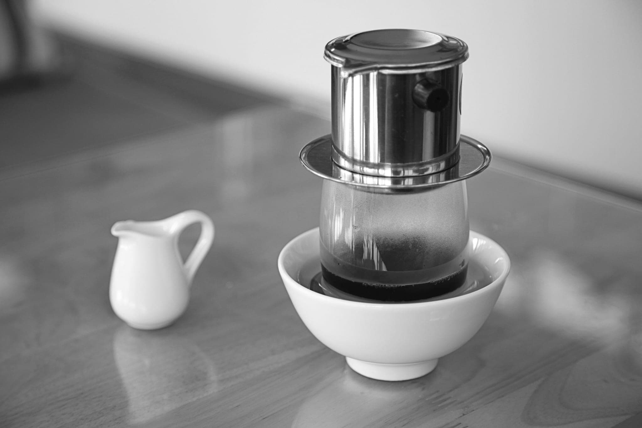 A stainless steel drip coffee filter on a glass set in a white bowl, beside a small milk jug on a wooden table