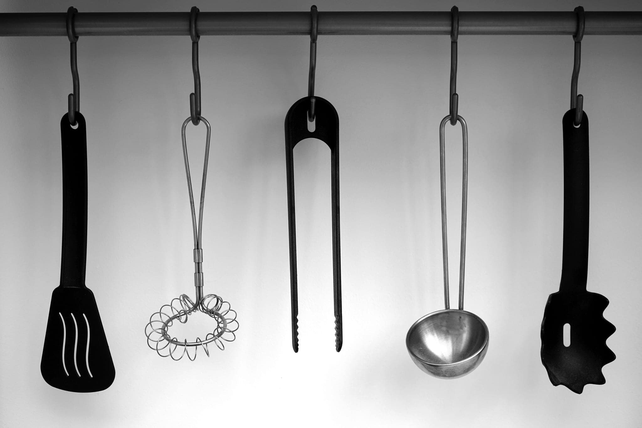 Five kitchen utensils hanging from a rail against a pale wall: a spatula, a whisk, tongs, a ladle and a pasta server