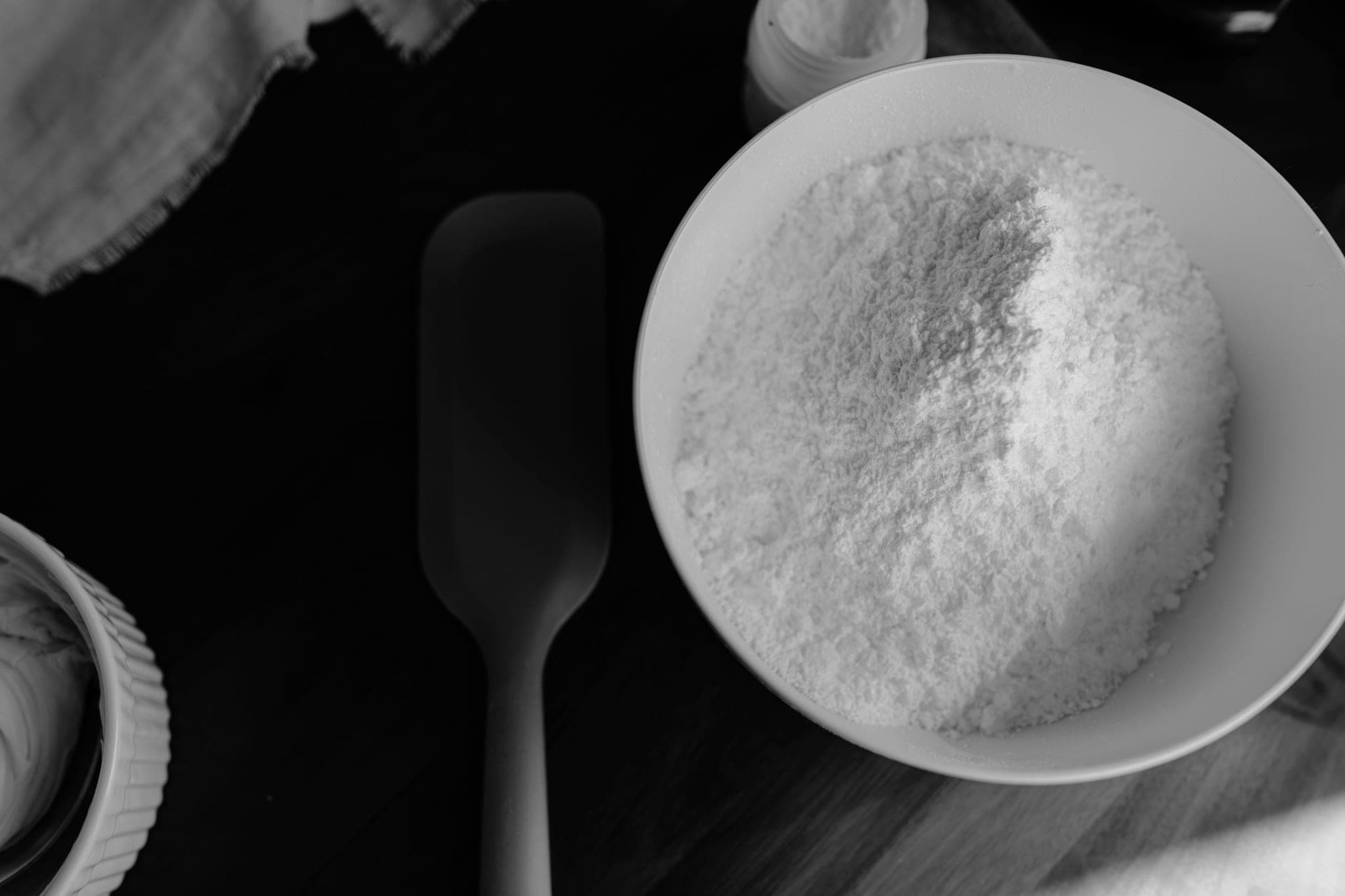 A white bowl heaped with fine white powder on a dark wooden table, beside a spatula