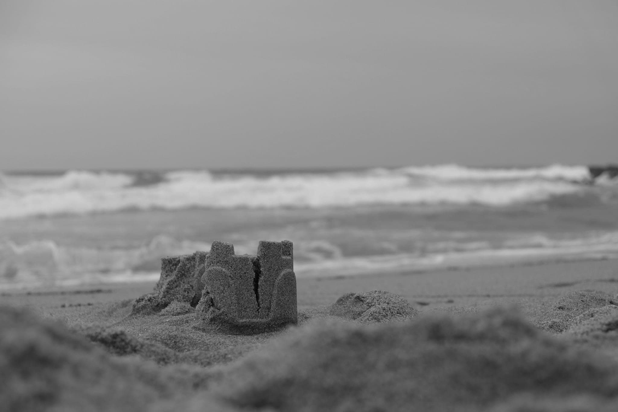 A small sandcastle on a beach, one tower already cracked, with waves breaking behind it