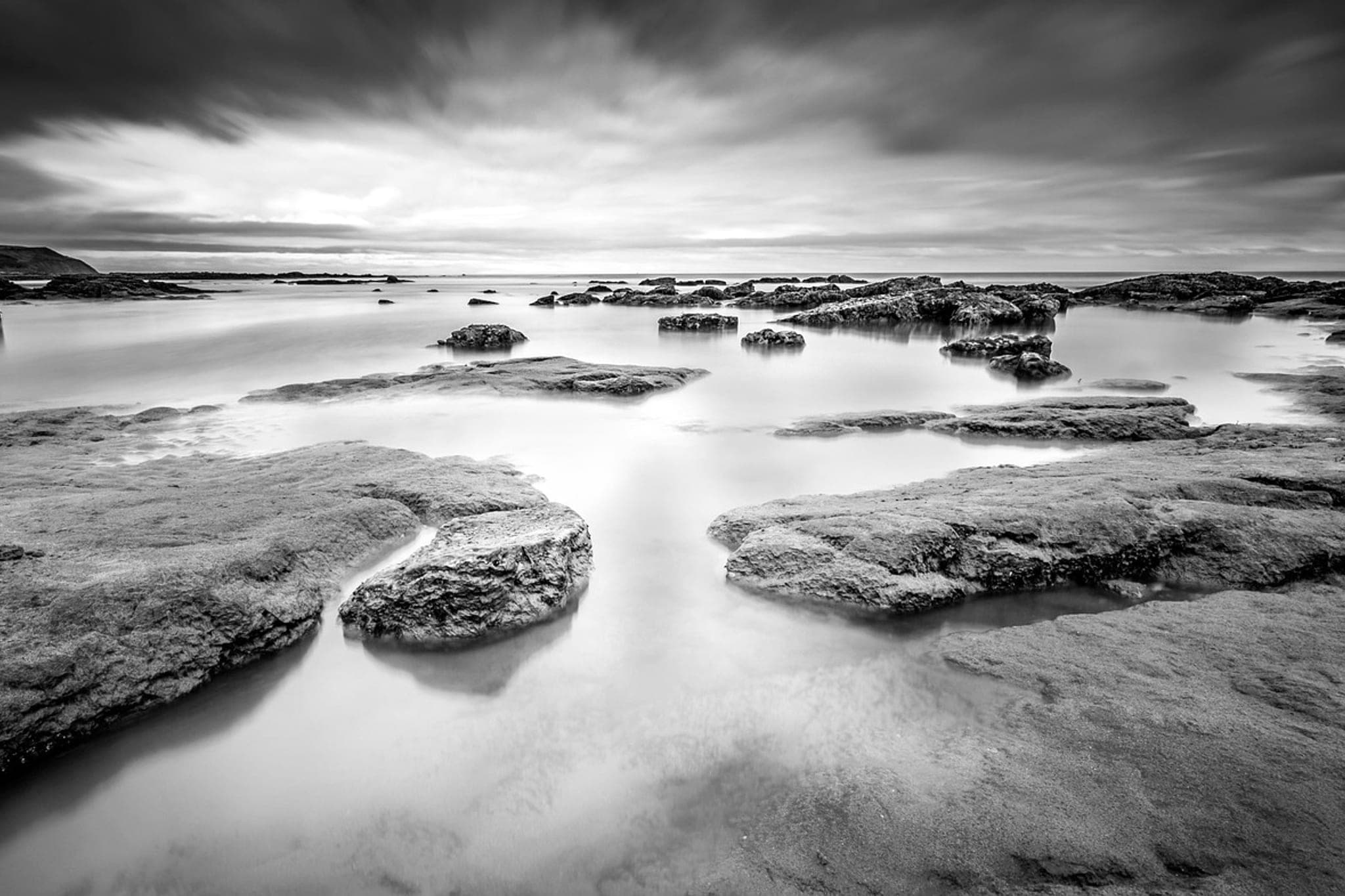 A rocky shore at low tide, water lying still between flat stone shelves under a heavy sky