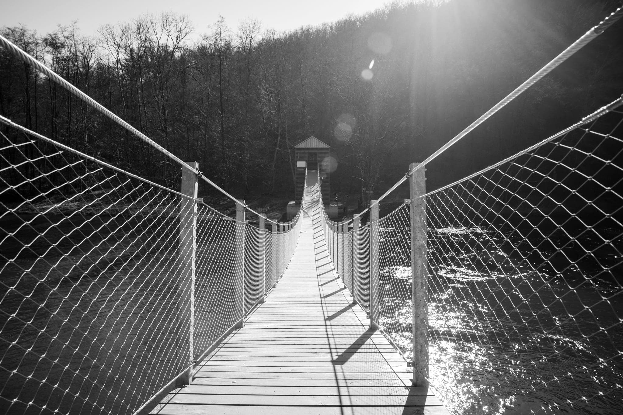A narrow suspension footbridge with mesh sides, crossing a river toward a small hut