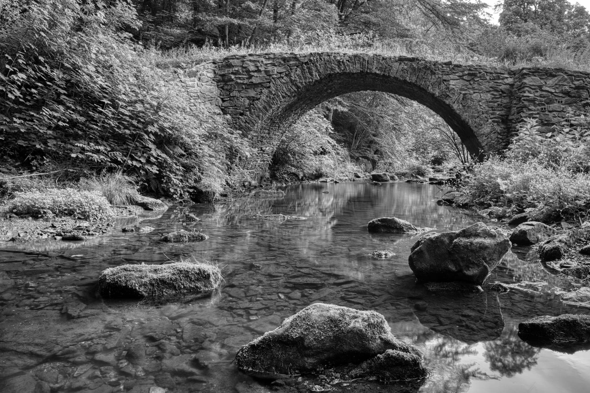 An old stone arch bridge over a shallow, rocky stream, reflected in the water