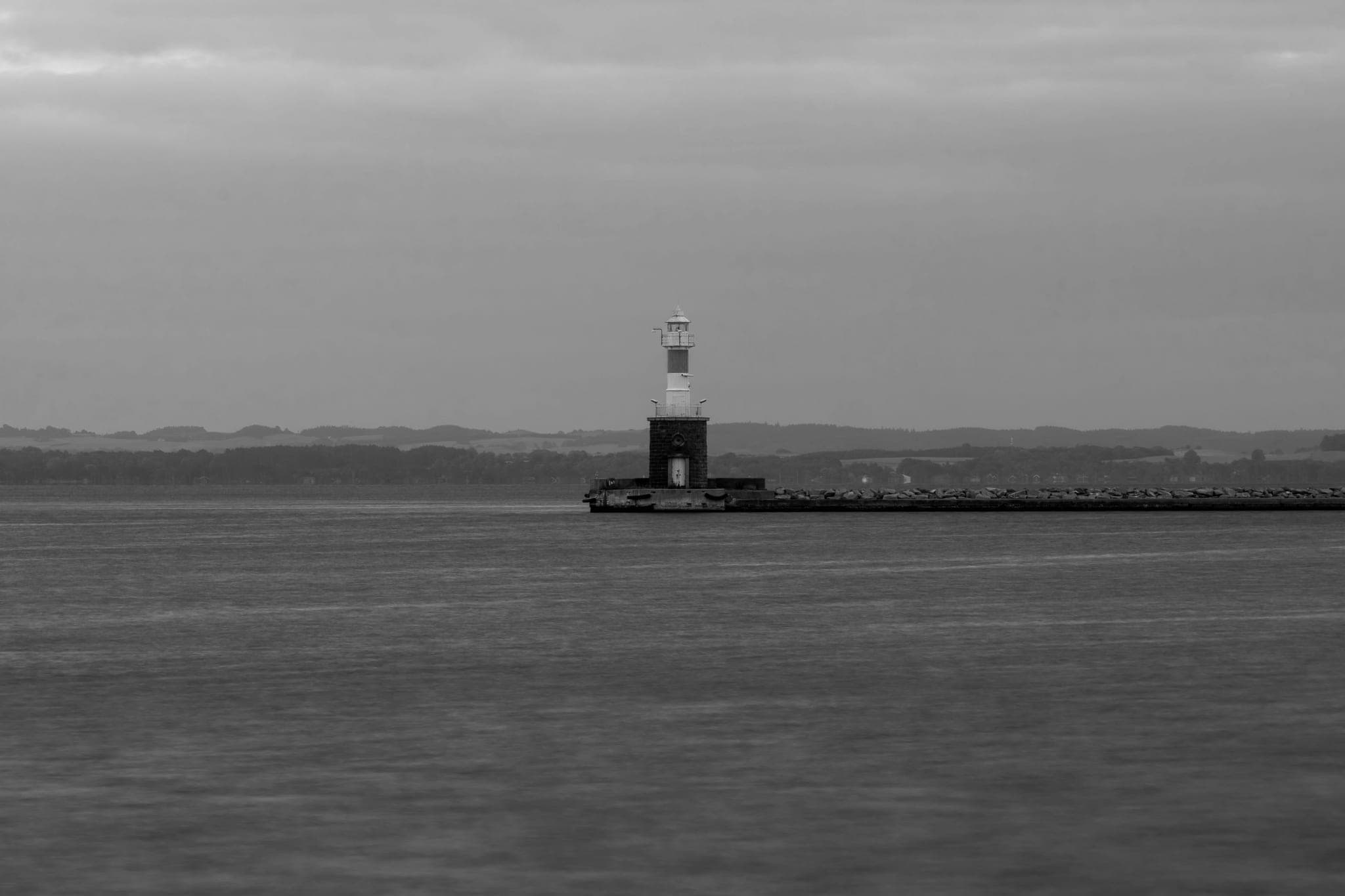 A small lighthouse at the end of a long breakwater, far across flat water