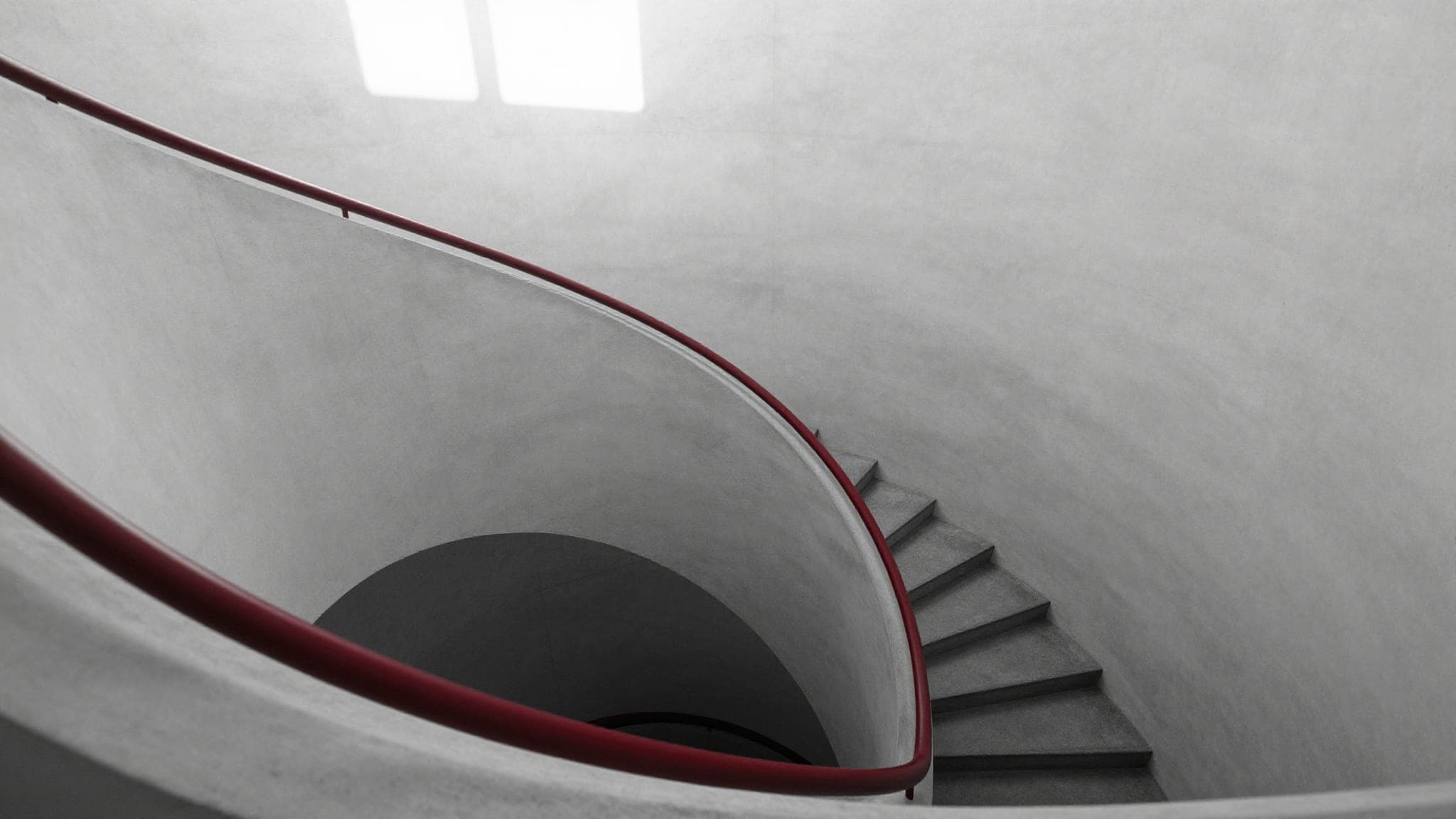 A concrete spiral staircase seen from above, one crimson handrail following the curve