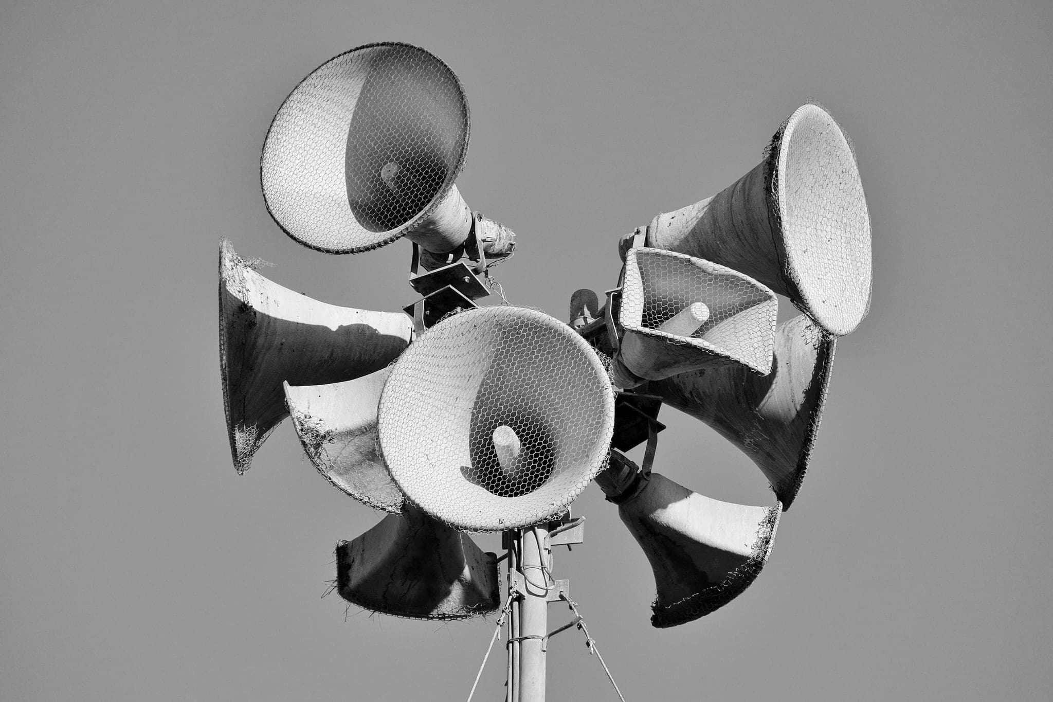 A cluster of weathered horn loudspeakers mounted on a pole, each pointing a different way against a plain sky