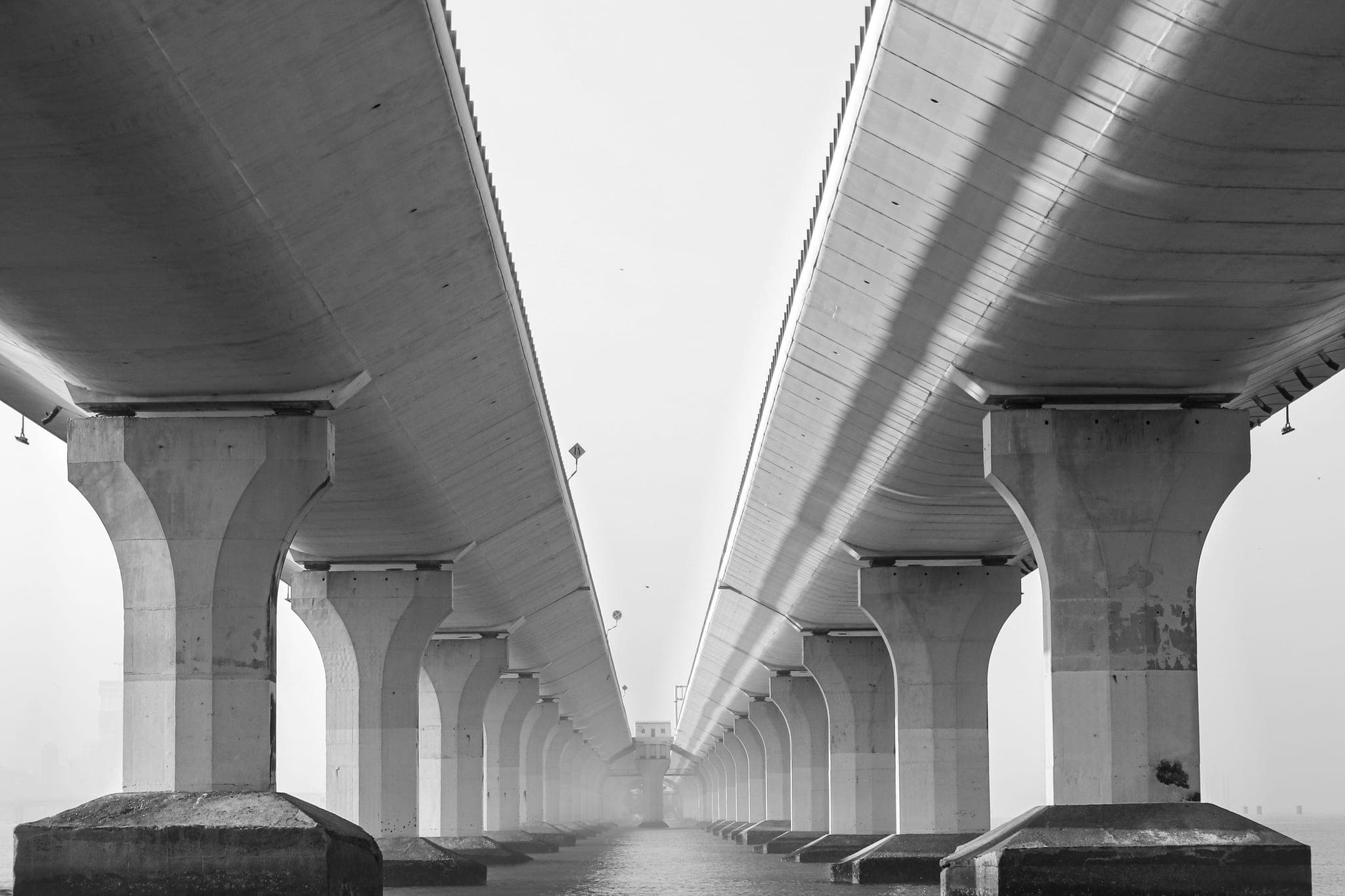 The undersides of two parallel bridge decks running side by side on concrete piers into the haze