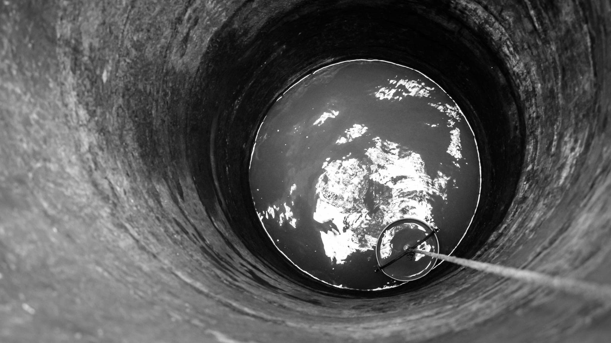 Looking down an old stone well at a bucket lowered on a rope toward the water